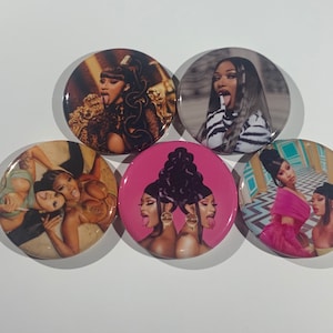 May include: A collection of five round buttons featuring images of female celebrities. The buttons display various portraits and poses, with a focus on vibrant colors and expressive imagery. The buttons are arranged on a white surface.