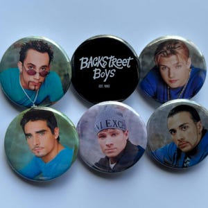 May include: A set of six circular pin badges. Five badges display images of men, and one is black with the white text "Backstreet Boys EST. 1993". The badges are arranged on a white background.