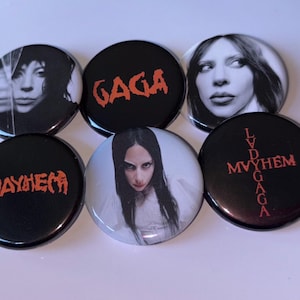 Op de afbeelding: Een verzameling van zes ronde knopen. Sommige knopen tonen zwart-wit portretten. Andere knopen hebben de woorden "GAGA", "MAYHEM" en "LVD GAGA MVYHEM" in rode, gestileerde tekst op een zwarte achtergrond.