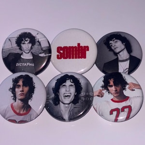 Könnte beinhalten: Sechs runde Ansteckbuttons. Fünf zeigen Schwarz-Weiß-Porträts einer Person mit lockigem Haar. Ein Button hat das Wort "sombr" in Rot. Die Buttons liegen auf einer weißen Oberfläche.