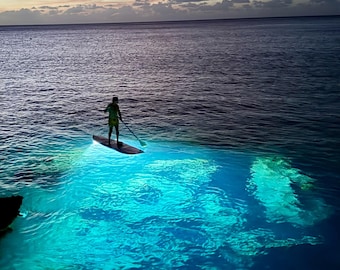 Könnte beinhalten: Eine Person paddelt auf einem Brett auf einem Gewässer bei Sonnenuntergang. Das Wasser hat eine leuchtend blaue Farbe und der Himmel ist in sanftem Rosa und Lila.