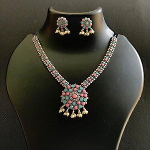 Puede incluir: Un conjunto de collar y pendientes plateados. El collar presenta un colgante con piedras rosas y turquesas, y los pendientes a juego tienen un diseño similar. Pequeñas cuentas blancas cuelgan del colgante y los pendientes.