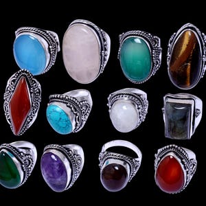 May include: A selection of silver rings with diverse gemstone settings. The rings display stones in blue, pink, green, brown, and purple hues. The gemstones are oval, round, and teardrop-shaped, set in ornate, detailed bands.