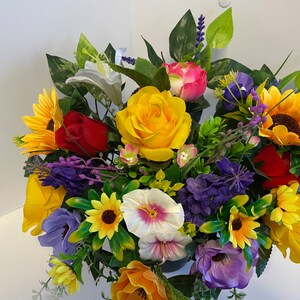 Könnte beinhalten: Ein bunter Strauß künstlicher Blumen in einer Vase. Der Strauß enthält Sonnenblumen, Rosen, Lavendel und andere Blumen in den Farben Gelb, Rot, Lila und Weiß.