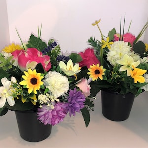 May include: Two black pots filled with colorful artificial flower arrangements. The arrangements include pink, yellow, white, and purple flowers.
