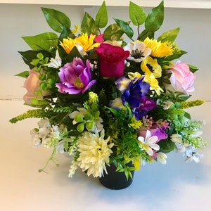 Può includere: Un arrangiamento di fiori artificiali colorati in un vaso nero. L'arrangiamento comprende fiori rosa, viola, gialli e bianchi con foglie verdi.