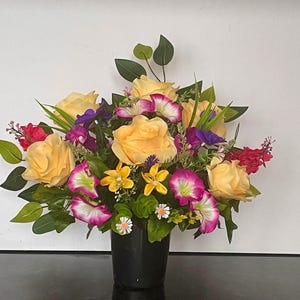 May include: A yellow, pink and purple artificial flower arrangement in a black vase. The flowers are arranged in a semi-circular shape and are surrounded by green leaves.