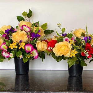 May include: Two artificial flower arrangements in black pots. Each arrangement features yellow roses, purple and pink flowers, and green foliage.
