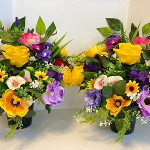May include: Two artificial flower arrangements in black pots. The arrangements feature a variety of flowers in shades of yellow, red, purple, and white.