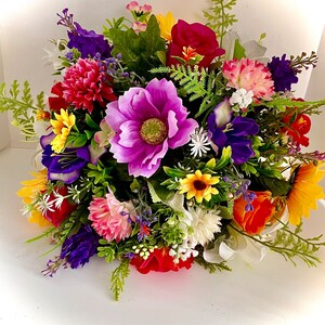 Pode incluir: Um buquê colorido de flores artificiais em tons de roxo, rosa, amarelo, vermelho e branco. O buquê apresenta uma variedade de tipos de flores, incluindo rosas, girassóis e margaridas.