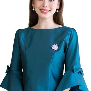 May include: A teal blouse with a boat neckline and three-quarter length bell sleeves featuring small bows at the cuffs. The garment is adorned with a silver-toned floral brooch on the left side of the chest, showcasing a structured, shimmering fabric.