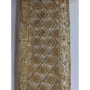Könnte beinhalten: Nahaufnahme eines goldfarbenen, verzierten Stoffes. Der Stoff weist ein sich wiederholendes Muster aus Rauten mit floralen Akzenten auf. Die Ränder sind mit einem detaillierten, dekorativen Design eingefasst. Der Stoff wird wahrscheinlich zur Verzierung oder Dekoration verwendet.