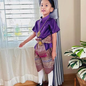 Könnte beinhalten: Ein Kind in traditioneller thailändischer Kleidung. Das Outfit besteht aus einem lila kurzärmeligen Hemd, gemusterten Shorts, einem dekorativen Gürtel und einer goldenen Halskette. Das Ensemble wird durch weiße Socken und schwarze Schuhe ergänzt.