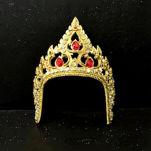 Op de afbeelding: Een goudkleurige kroon met rode en heldere edelstenen. De kroon heeft een centrale piek en een gebogen basis, met sierlijke details en een symmetrisch ontwerp. De edelstenen zijn in een decoratief patroon geplaatst.