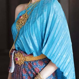 May include: A woman wearing a traditional Thai outfit. The ensemble features a light blue pleated top, a patterned skirt, and gold jewelry, including a necklace, earrings, bracelets, and a belt with a large central ornament.