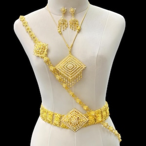 Pode incluir: Um conjunto de joias douradas exposto em um manequim. O conjunto inclui um colar com um pingente em forma de diamante, brincos combinando e um cinto. As joias são intrincadamente projetadas com padrões florais e geométricos e pequenos detalhes pendurados.