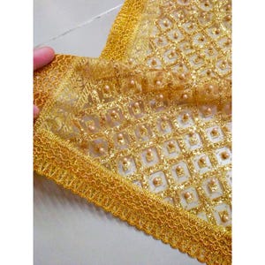 Puede incluir: Primer plano de una tela dorada y ornamentada con un patrón cuadrado repetido. La tela está adornada con un borde de encaje dorado decorativo. Pequeñas cuentas, similares a perlas, están intercaladas en el diseño, agregando textura y dimensión.