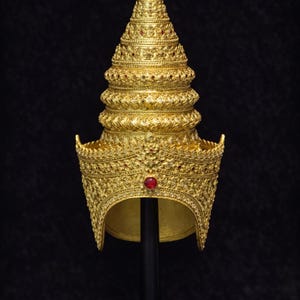 Può includere: Una corona dorata ornata, con una parte superiore conica e dettagli intricati. La corona presenta accenti di pietre preziose rosse ed è esposta su un supporto nero su sfondo scuro. Tema cerimoniale o storico.