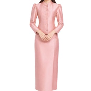 Könnte beinhalten: Ein hellrosa formeller Anzug, bestehend aus einer langärmeligen Jacke mit Knopfverschlüssen und einem passenden bodenlangen Rock. Die Jacke hat einen Stehkragen und Puffärmel. Das Outfit wird mit nudefarbenen Absätzen kombiniert.