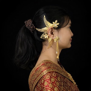 May include: A side profile of a person wearing ornate gold ear jewelry. The ear cuff features a floral and wing design, while the earring has a long chain with a pearl. The person is wearing a red and gold patterned garment.