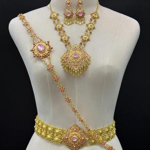 May include: A gold-toned jewelry set featuring a necklace, matching drop earrings, and a decorative waist belt. Each piece is crafted with intricate filigree metalwork, pink oval-shaped center stones, and small pink accents throughout the design.
