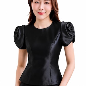 Op de afbeelding: Een zwarte blouse met korte mouwen, een ronde halslijn en een gladde, satijnachtige afwerking. De mouwen hebben een opvallend geplooid ontwerp dat op elke schouder een roosachtige vorm vormt, en het lijfje is getailleerd met verticale naden.
