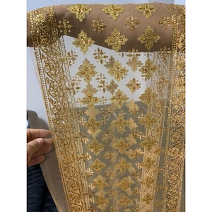 Puede incluir: Una tela transparente de color beige con intrincados bordados dorados. El diseño presenta patrones florales y bordes repetidos, creando una apariencia lujosa y ornamentada. La tela está drapeada, mostrando su delicada textura y detalles brillantes. (Aprox. 100 cm)