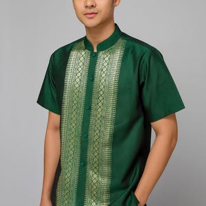 Può includere: Una camicia verde scuro a maniche corte con colletto alla coreana e un motivo dorato sul davanti. La camicia ha una tasca sul lato sinistro ed è abbottonata. La camicia è indossata da una persona.