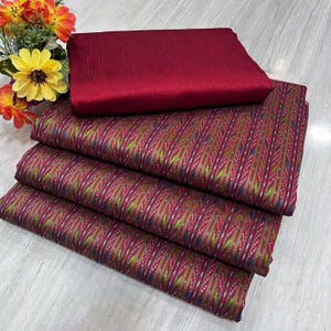 May include: A stack of patterned fabric in shades of burgundy, green, and blue, with a solid burgundy piece on top. The fabric has a textured appearance and is likely intended for sewing or crafting projects. A bouquet of flowers is in the background.