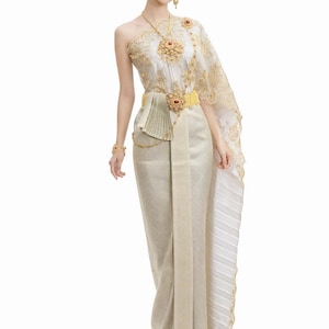 May include: A traditional Thai dress in cream and gold. The one-shoulder top and skirt are adorned with intricate gold embroidery and embellishments. A gold belt with a red jewel accentuates the waist. The ensemble includes a matching headpiece and jewelry.