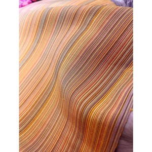 Puede incluir: Primer plano de una tela con un patrón de rayas. La tela presenta una mezcla de colores cálidos, incluyendo naranja, amarillo, rojo y marrón, dispuestos en rayas verticales. La tela parece estar tejida, con una superficie ligeramente texturizada.