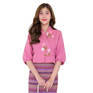 Puede incluir: Una blusa rosa con mangas tres cuartos y cuello cruzado. La blusa presenta bordados florales en blanco, rosa y verde. La modelo lleva una falda a rayas con rayas moradas, marrones y naranjas.