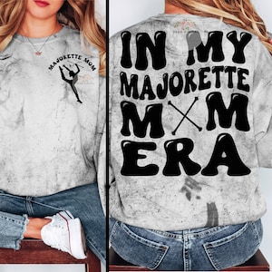 May include: A gray sweatshirt with a marble-like pattern. The front features the text "MAJORETTE MOM" and a silhouette of a majorette. The back displays "IN MY MAJORETTE MXM ERA" in large, black, bubble letters. A casual, comfortable top.