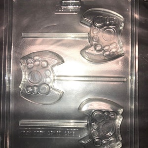 May include: A clear plastic mould for making chocolate game controllers. The mould has two cavities, each shaped like a game controller with buttons and a directional pad. The mould is designed to be used with a lollipop stick.