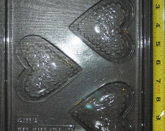 May include: A clear plastic mould for making chocolate hearts. The mould has three heart-shaped cavities, each with a detailed design. The mould is labelled "Copyright 1998 W.E."