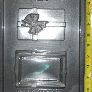 May include: A black plastic mold with two cavities. The top cavity is shaped like a gift box with a bow. The bottom cavity is shaped like a rectangular picture frame. The mold is labeled "East Coast Pro Mold, Inc. 401 855"