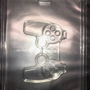 May include: A clear plastic mould for making chocolate video game controllers. The mould is shaped like a video game controller with buttons and a directional pad. The mould is in a clear plastic tray with the text "AO 1851" and "BUSCH  CHOCOLATE MOLD  MFG  USA" printed on it.