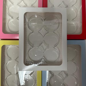 Op de afbeelding: Een verzameling kleurrijke cupcake-dozen in roze, rood, geel en blauw. Elke doos bevat een doorzichtige plastic inzet met zes ronde compartimenten. De dozen zijn gerangschikt met een witte doos erboven.