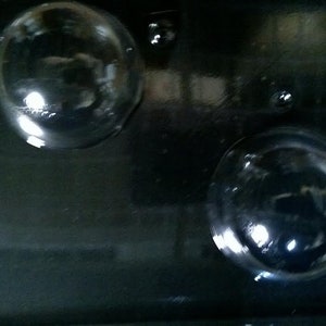 May include: Two clear, spherical glass objects are set against a dark, reflective surface. The spheres appear to be lenses or decorative elements, possibly part of a larger device or artwork. The background is a dark, possibly metallic, material.