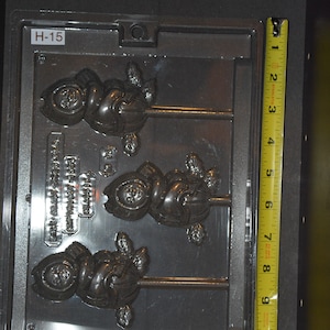 May include: A gray plastic mold with three cavities for making lollipops. Each cavity features a detailed design of a cartoon character wearing a hat and holding a ball. The mold is labeled "H-15" and "CLASSIC SANTA & ELF POP"