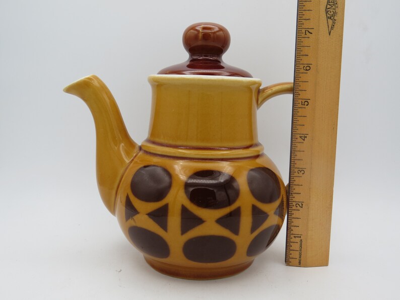 May include: A brown and yellow ceramic teapot with a brown lid and a geometric pattern of brown circles and triangles. The teapot is about 6 inches tall.
