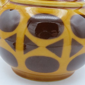 May include: A yellow teapot with a brown geometric pattern. The teapot has a round body and a spout. The pattern consists of circles and triangles.