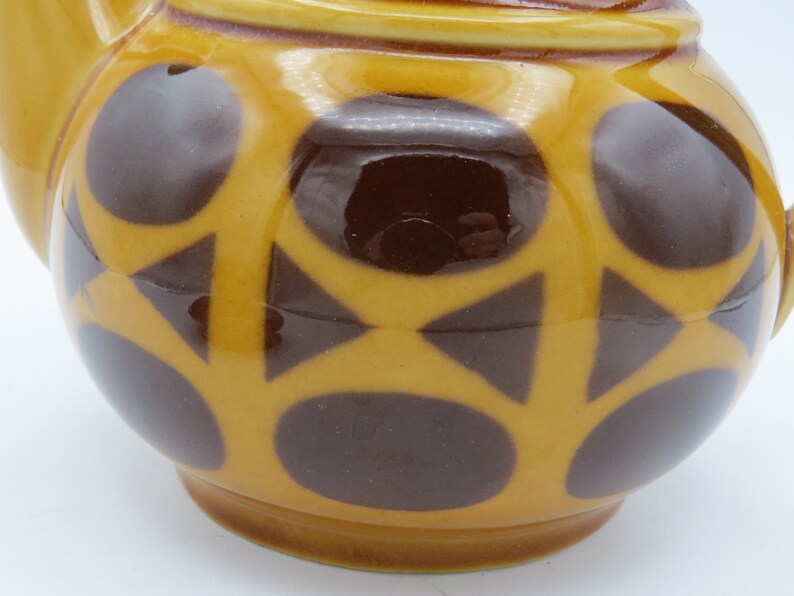 May include: A yellow teapot with a brown geometric pattern. The teapot has a round body and a spout. The pattern consists of circles and triangles.