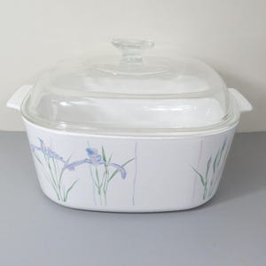 May include: White casserole dish with a clear glass lid. The dish has a floral design with purple irises and green leaves. The dish has two handles and is suitable for baking and serving.