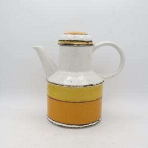 May include: A ceramic coffee pot with a white body and a yellow and orange base. The pot has a curved handle and a spout. The lid is yellow with brown trim. The pot is approximately 9 inches tall.