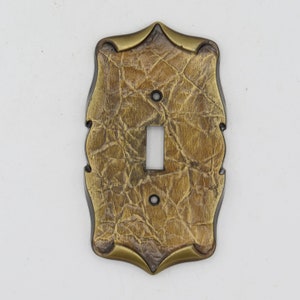 Pode incluir: Uma tampa de interruptor de luz decorativa com um design texturizado que imita a pele de crocodilo. A tampa é dourada e marrom com um contorno preto e uma única abertura para o interruptor.
