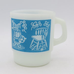 Op de afbeelding: Witte keramische mok met een blauwe band waarop een wit silhouet van een stoel en een schommelstoel is afgebeeld. De mok heeft een afgeronde handgreep.