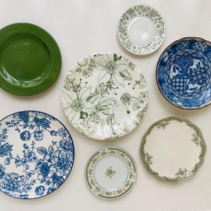 May include: A collection of seven ceramic plates featuring various designs: one solid green plate, one with green botanical illustrations, one with blue and white floral patterns, one with blue and white geometric motifs, and three smaller plates with floral borders.