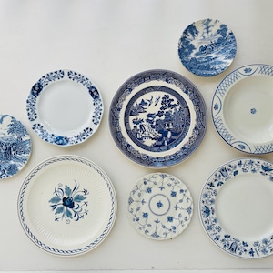 May include: A collection of eight ceramic plates featuring various blue and white patterns. The set includes plates with landscape scenes, floral motifs, geometric borders, and intricate willow designs, all arranged in a flat, top-down view.