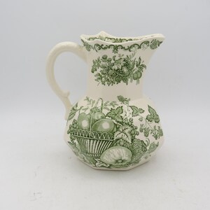 May include: Cream ceramic jug with a green fruit basket and floral pattern. The jug has a curved handle and a scalloped rim. The fruit basket design includes apples, pears, and grapes. The jug is likely for serving beverages, such as water or juice.